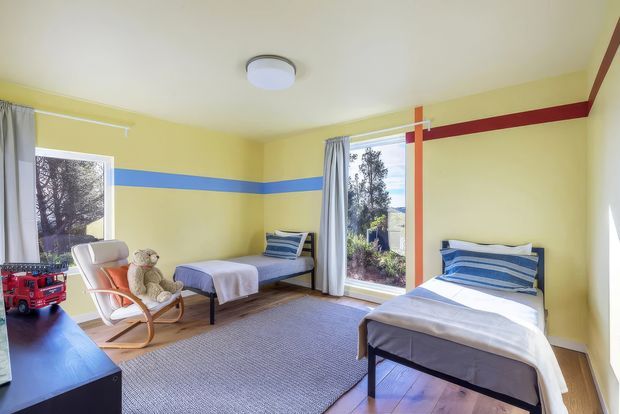 A bright shared bedroom with two single beds, yellow walls accented by horizontal blue and red stripes, timber flooring, a large grey rug, and full-height windows with curtains. A rocking chair and teddy bear sit beside a dark dresser, creating a playful yet tidy children's room.
