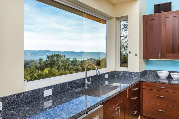 Bright contemporary kitchen with dark polished granite benchtops, warm wood cabinetry, stainless-steel sink and tapware, and expansive windows overlooking a wooded landscape and distant hills. A vivid blue wall section adds colour contrast.