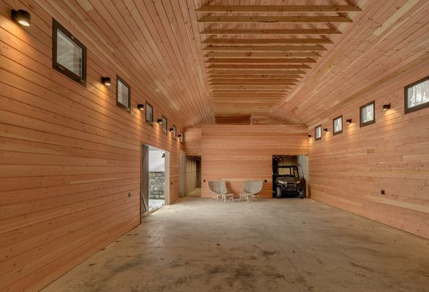 A large, elongated timber-lined interior with horizontal wood wall boards and a matching plank ceiling. Exposed timber rafters run along the central ceiling, while small high-level windows and wall-mounted lights line both sides. The unfinished concrete floor, open plan, dining table and chairs, and dark storage or equipment area suggest a spacious barn-like workshop, studio, or garage interior.