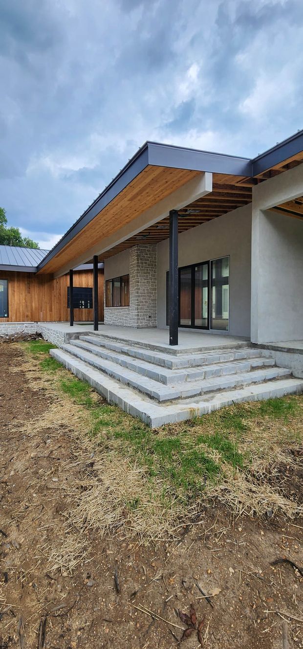 Exterior view of a contemporary low-rise building with broad flat roof overhangs, exposed timber soffits and rafters, dark slender steel columns, natural timber cladding, pale rendered walls, and a grey stone feature wall. Wide masonry steps lead up to glazed entrance and living areas. The surrounding ground is unfinished, with grass and soil in the foreground beneath an overcast sky.