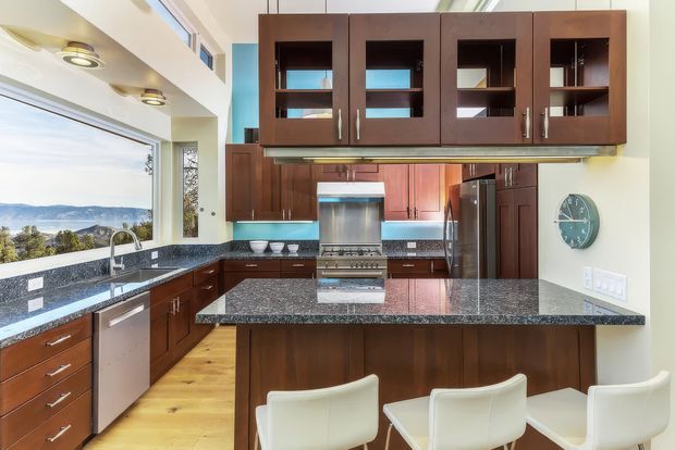 Bright contemporary kitchen with dark wood cabinetry, extensive granite countertops, a large central island with seating for three, stainless steel appliances, and light timber flooring. Large windows provide an expansive landscape view, while a turquoise accent wall and clerestory glazing add colour and light.