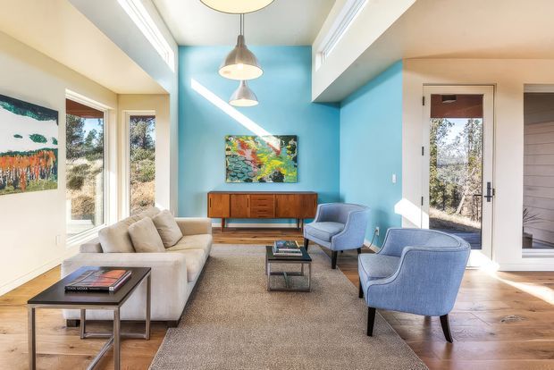 A bright, colourful mid-century-inspired living room with a vivid turquoise feature wall, warm timber flooring, a cream sectional sofa, blue upholstered lounge chairs, a low timber sideboard, and a central coffee table. Large windows and glazed doors provide abundant natural light, while cone pendant lights, artwork, and layered ceiling detailing add visual interest.