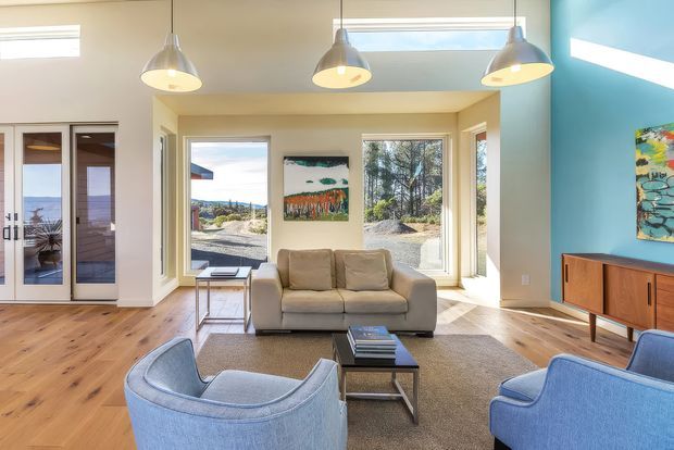 Bright contemporary living room with a beige sofa, two blue upholstered lounge chairs, a dark coffee table, and a timber media console. The room has warm timber flooring, pale walls, a vivid blue accent wall, large windows, glazed double doors, and three suspended pendant lights. Outdoor landscape is visible through the glazing.