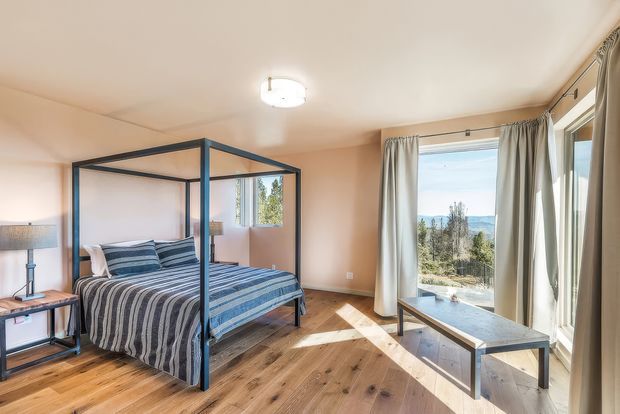 Bright bedroom with a dark-framed four-poster bed dressed in blue and grey striped bedding, warm timber flooring, pale peach walls, and full-height curtained glazing. A simple coffee table and side table sit in the foreground, with a scenic outdoor view visible through the windows.