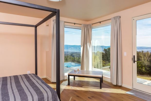 Bright bedroom with a four-poster bed, striped bedding, warm timber flooring, pale peach walls, and floor-to-ceiling corner glazing overlooking a landscaped outdoor area and distant mountains. A small dark timber coffee table sits beside the windows, with full-height curtains and a glazed exterior door.