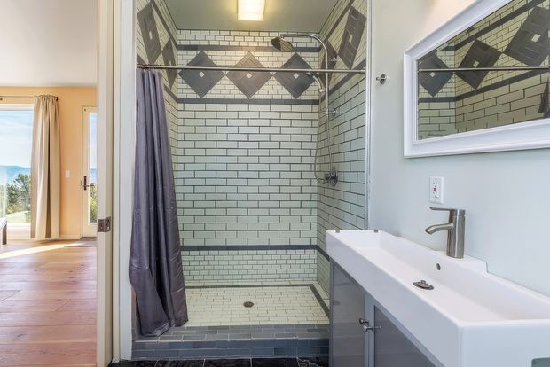 Bright contemporary bathroom with a walk-in tiled shower featuring pale subway tiles, dark grout, and a decorative geometric tile band. A large wall mirror, white vanity basin, chrome tapware, and grey cabinetry occupy the foreground. An adjacent timber-floored room is visible through the open doorway.
