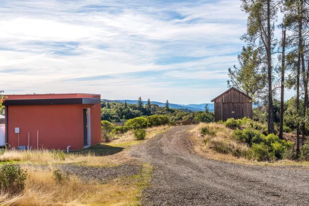 A rural property setting with a compact modern red outbuilding on the left, a weathered timber shed or barn on the right, and a broad gravel driveway leading through dry grasses and shrubs toward rolling wooded hills. Tall trees frame the scene under a bright, lightly clouded sky.