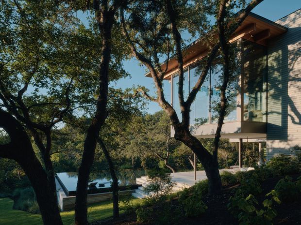 Exterior view of a contemporary multi-level residence set among mature trees, with pale horizontal cladding, extensive floor-to-ceiling glazing, projecting timber roof eaves, and an elevated terrace or balcony. A swimming pool and landscaped garden are visible below, integrated into the wooded site.