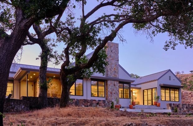 Single-storey contemporary home set in a dry, rocky landscape, photographed at dusk. The building has multiple pitched metal-roof volumes, extensive dark-framed glazing, pale rendered walls, and prominent rough natural-stone cladding around a tall central chimney and low base walls. Mature trees frame the facade, with warm interior lighting visible through the windows and a covered entry terrace on the left.