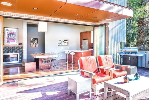 Bright modern indoor-outdoor living area with an open glazed wall connecting a timber deck to a compact kitchen and dining space. The room features a vivid orange soffit, corrugated metal exterior wall, polished timber flooring and decking, red upholstered lounge chairs, a compact kitchen island, a dining table, and an outdoor barbecue. A patterned tiled fireplace surround is visible at the left.
