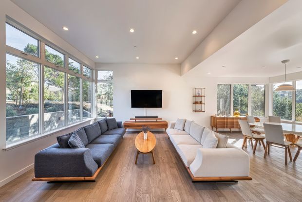 Bright, open-plan contemporary living and dining area with floor-to-ceiling glazing, white walls, recessed lighting, warm timber flooring, two large sofas, a wall-mounted television, and a timber dining table near the windows.