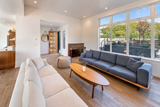 Bright contemporary living room with large white-framed windows, warm timber flooring, two sofas, an oval timber coffee table, and a compact timber console or piano. An open-plan kitchen and entry area are visible at the left and rear, with a planted outdoor area seen through the glazing.