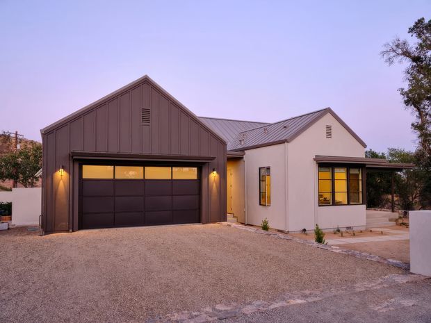 Exterior view of a single-storey residential property at dusk, featuring a prominent dark metal-clad gabled garage, attached light-rendered living wing, pitched metal roofs, warm illuminated windows, and a broad gravel driveway.