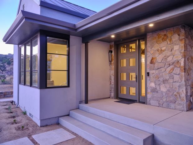 Modern residential house exterior focused on a covered front entry. The facade combines smooth light-grey render, dark metal-framed glazing and doors, a low-pitched metal roof, and a prominent irregular natural-stone-clad wall. Broad concrete steps lead to a recessed porch with recessed downlights. Sparse landscaped ground and distant terrain are visible around the entry.