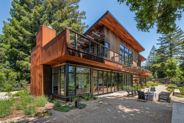 Exterior view of a multi-level residential home surrounded by mature trees, featuring a prominent pitched gable roof, warm timber siding and soffits, dark metal-framed floor-to-ceiling glazing, and projecting upper-level balconies. A paved outdoor seating terrace with lounge chairs occupies the foreground.