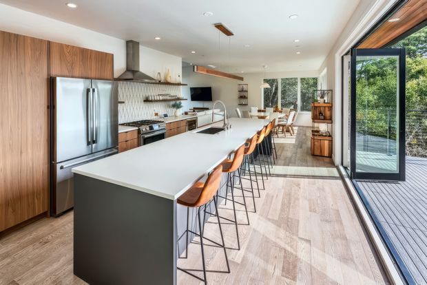 Bright, spacious contemporary kitchen with a long white island and multiple bar stools, full-height timber cabinetry, stainless-steel appliances, patterned tile splashback, open shelving, and large glazed doors opening onto a timber deck. The kitchen extends toward a dining and living area with abundant natural light and greenery beyond.