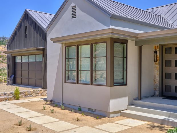 A sunlit residential house exterior with a pale rendered facade, prominent projecting bay window with dark timber-look frames, and a covered entry porch. The home has light grey metal gable roofs, a dark grey attached garage, stone-clad entry detailing, concrete stepping paths, and a dry landscaped frontage.