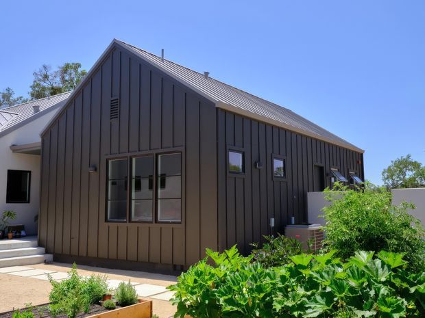 Exterior view of a contemporary single-storey gabled building clad in dark vertical metal panels, with a pitched metal roof, black-framed windows, an adjoining lighter wing, paved entry steps, and a productive garden in the foreground.