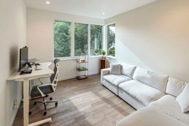 Bright, compact multipurpose room with a white sectional sofa, a minimalist desk and ergonomic office chair, open shelving, and large corner windows overlooking greenery. The room has white walls, recessed ceiling lights, and light wood-look flooring, creating a clean contemporary work-and-lounge setting.
