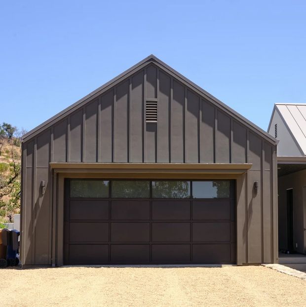 Front exterior view of a dark charcoal-brown detached garage or outbuilding with a prominent pitched gable roof, vertical standing-seam style wall panels, and a wide sectional garage door with upper glazed panels. A neighbouring pale metal-clad structure is partially visible at right, with a gravel driveway in front.