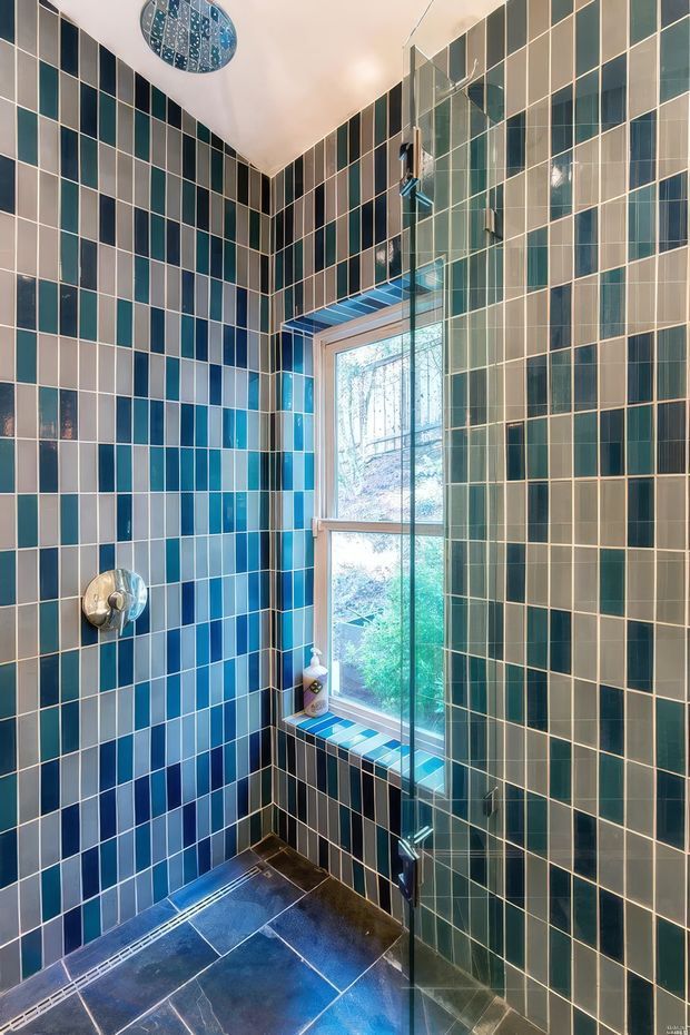 A compact contemporary shower room lined floor-to-ceiling with a geometric grid of glossy rectangular tiles in deep blue, teal, grey, and white. A frameless glass shower screen encloses the shower, with a window providing natural light and views of greenery. The floor is dark tiled, and a patterned blue ceiling light adds a decorative accent.