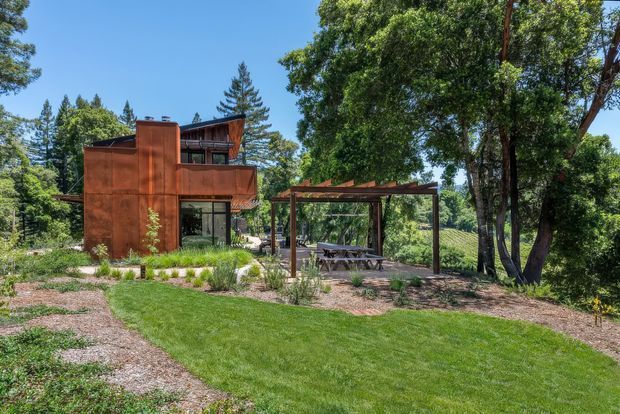 Exterior view of a modern, multi-level house set within a mature wooded landscape. The home features a rust-toned metal-clad vertical volume, extensive glazing, dark timber balcony and roof elements, and an adjacent timber pergola over an outdoor dining area. A landscaped lawn, gravel paths, ornamental planting, and established trees surround the building.