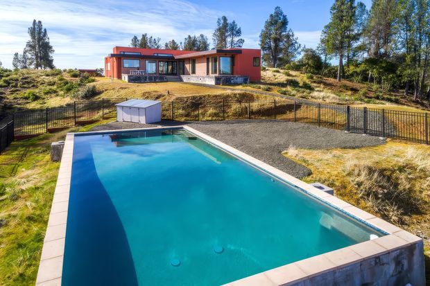 A modern single-storey red-clad home set on a landscaped, sloping site, viewed from a large rectangular swimming pool in the foreground. The property features dark-framed glazing, a low-profile roofline, gravel and grass landscaping, and a black metal boundary fence surrounded by mature trees.