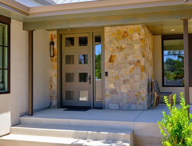A bright residential front entry viewed from outside, featuring a covered porch with broad concrete steps, a contemporary dark-framed glazed entry door, large windows, and a prominent irregular natural-stone-clad wall. Timber-toned posts and a layered flat roof or clerestory-style canopy frame the entrance, with a small garden shrub at the edge.