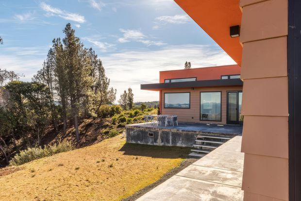 Exterior view of a contemporary single-storey house on a landscaped, sloping site. The foreground shows a concrete walkway and terracotta-toned wall with dark vertical cladding, while the main building has horizontal timber-toned cladding, broad glazing, a projecting flat roof and a raised concrete terrace with outdoor dining furniture and steps. Mature trees and distant hills surround the property.