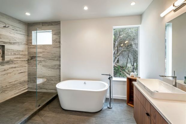 Bright contemporary bathroom featuring a freestanding white bathtub, walk-in glass shower, double vanity with timber cabinetry and white benchtop, large-format stone-look wall and floor finishes, recessed ceiling lights, and a window overlooking trees.
