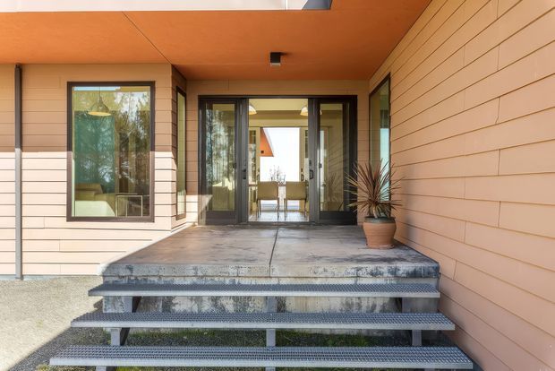 A contemporary residential entry viewed from outside, with broad concrete steps and metal-grate treads leading to a covered porch. The facade is clad in horizontal light timber-toned boards, complemented by a warm wood soffit and dark-framed glazing. A glazed double entry door reveals a bright interior beyond, while a potted architectural plant adds a small accent beside the doorway.