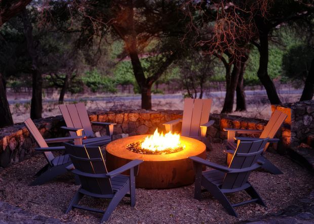 A landscaped outdoor seating area with several dark timber lounge chairs arranged around a circular fire pit. A low natural-stone retaining wall encloses the space, with mature trees and dense vegetation surrounding the intimate gathering area. The fire provides a warm focal glow against the moody dusk setting.