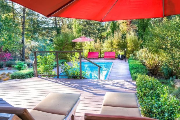A landscaped outdoor pool area viewed from a timber deck beneath a large red shade umbrella. The scene includes a bright blue swimming pool, timber decking and walkway, glass-and-timber balustrade, vivid pink outdoor seating and umbrella, and dense mature garden planting.