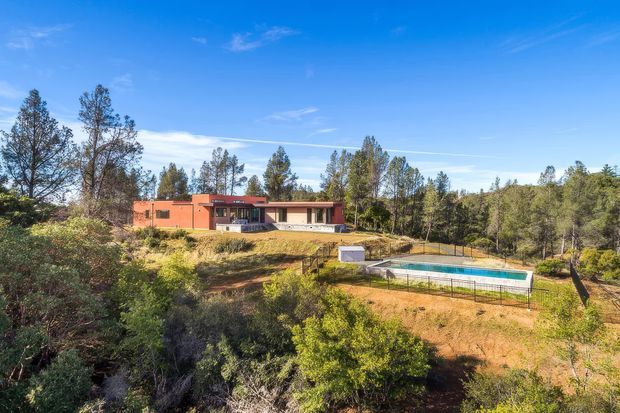 Wide exterior view of a low, single-storey contemporary home set within a dry, wooded landscape. The building has long horizontal proportions, terracotta-coloured rendered walls, dark-framed glazing and a shallow roofline. A fenced rectangular swimming pool occupies the foreground, with native-looking vegetation and mature trees surrounding the property under a clear blue sky.