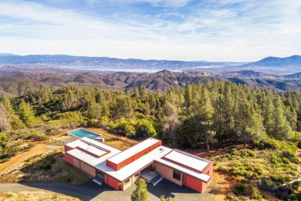 Aerial view of a low-slung, modern single-storey residence with bold red and white exterior walls, flat roof forms, and a small turquoise swimming pool. The house is set among dense trees and rugged, expansive mountainous terrain, with a bright open sky and dramatic panoramic landscape beyond.