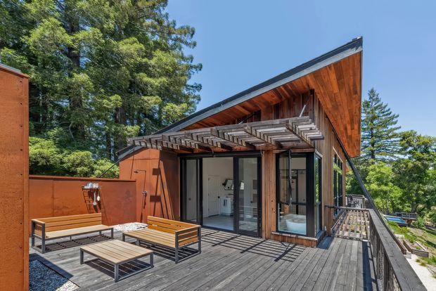 Exterior view of a modern timber-clad house with a dramatic sloping roof and exposed timber soffit. A large timber deck with black metal-framed outdoor benches and a low coffee table extends around the building, while floor-to-ceiling glazing opens to the interior. The property is set among mature trees and appears elevated above the surrounding landscape.