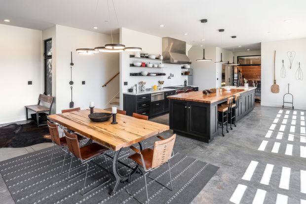 A spacious open-plan kitchen and dining area with a large timber dining table, leather-and-metal dining chairs, a black kitchen island, open shelving, dark cabinetry, stainless steel appliances, and polished concrete flooring. Pendant lights hang above the dining table and island, while strong sunlight creates geometric patterns across the floor. Decorative wall-mounted objects and a staircase are visible in the background.