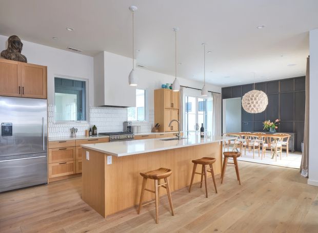 Bright, open-plan contemporary kitchen and dining area with a large light-wood island, white stone benchtop, timber cabinetry, stainless-steel appliances, white textured tile splashback, and three timber stools. A dining table with woven chairs sits beyond the island, with dark built-in cabinetry along the rear wall and a sculptural pendant light above.