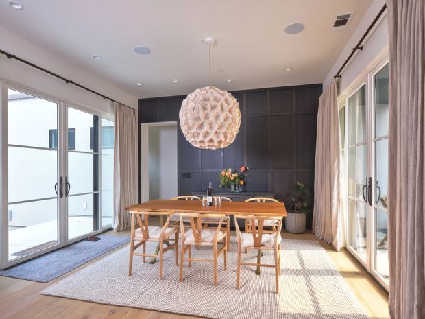 Bright, refined dining room with a long timber dining table and matching chairs, large glazed French-style doors on two sides, full-height curtains, a dark charcoal panelled wall with integrated cabinetry, and a sculptural woven pendant light. Natural light creates a warm, airy atmosphere, with a neutral rug and timber flooring grounding the space.