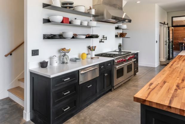 A spacious industrial-style kitchen with dark charcoal cabinetry, a white benchtop, stainless-steel range and oversized extraction hood, open black metal shelving displaying crockery, and a large timber island in the foreground. Concrete-look flooring and a visible stairway contribute to the utilitarian character, while an adjoining hallway and timber-clad area are visible beyond the kitchen.