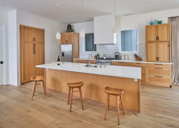 Bright open-plan kitchen featuring a large light timber island with white benchtop and integrated sink, matching timber cabinetry, white walls, pale timber flooring, stainless-steel appliances, and three timber stools. A tiled splashback, pendant lights, and windows complete the contemporary space.
