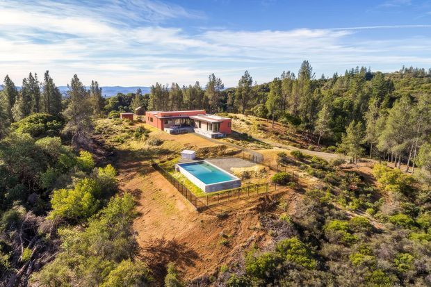 Aerial view of a low, modern single-storey residence set within a spacious wooded and scrub-covered rural landscape. The home has a long, low profile with a warm red exterior, broad glazing and a flat or very low-pitched roof. A rectangular swimming pool with a pale surround sits in the foreground, with informal paths and fencing around the property. Rolling hills and dense trees extend into the background beneath a bright blue sky.