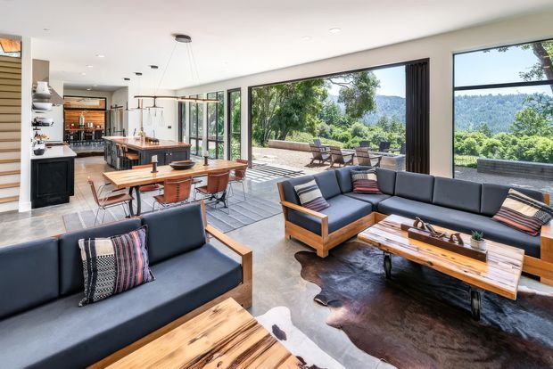 A bright, open-plan contemporary living and dining area connected to a modern kitchen. The room features extensive floor-to-ceiling glazing opening onto an outdoor terrace, dark leather sofas with timber frames, a timber coffee table, a dining table with bench seating, pendant lights, and views of a green wooded landscape. The interior combines warm natural timber, black accents, and polished concrete-style flooring.