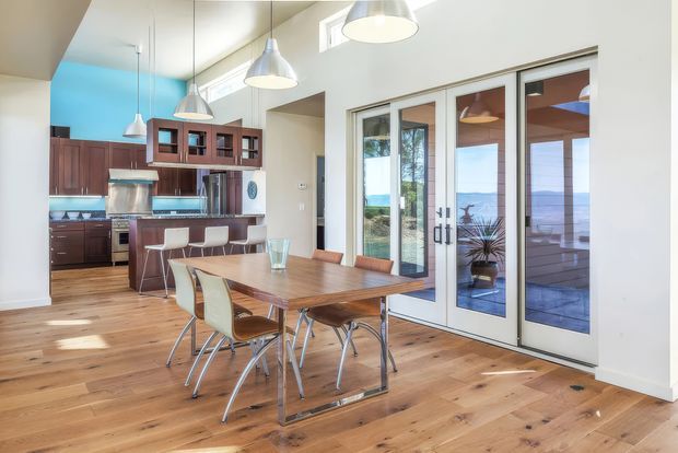 Bright open-plan kitchen and dining area with warm timber flooring, dark wood kitchen cabinetry, a peninsula with bar seating, and a rectangular timber dining table. Large glazed French-style doors open toward a deck with a visible coastal water view. Pendant lights, high ceilings, and clerestory windows create an airy contemporary interior.