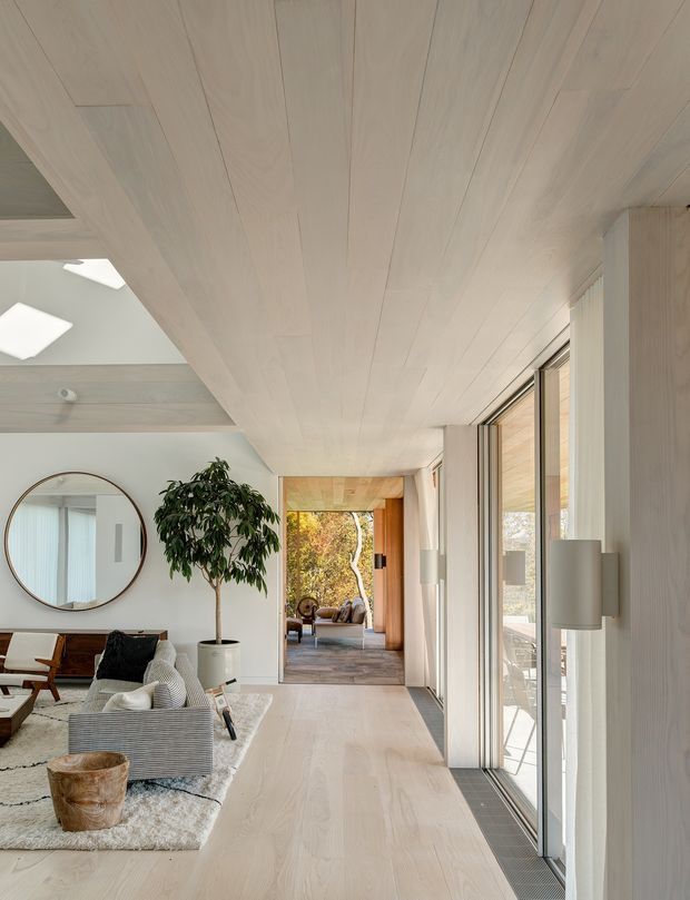 A bright, refined modern living room and circulation space with pale timber flooring and matching timber ceiling boards. Full-height glazing runs along the right side, bringing in abundant daylight and opening toward an outdoor terrace. The room contains a light neutral sofa, textured rug, low timber furniture, a round wall mirror, indoor tree, and sculptural lamp. A wide opening leads to another furnished sitting area, creating a strong sense of visual connection and depth.