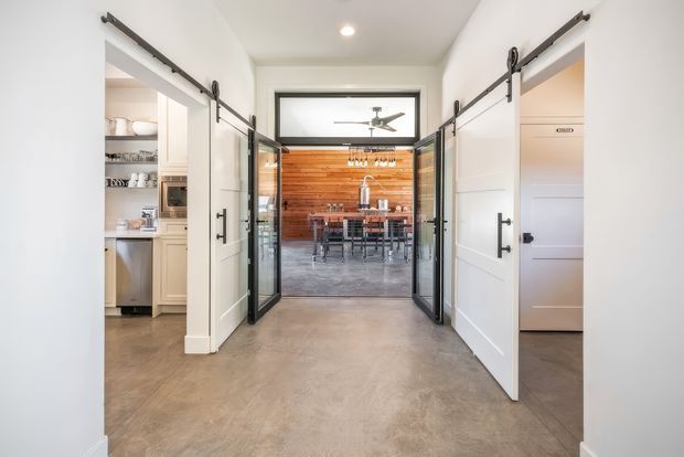 A bright modern interior corridor with polished concrete flooring and white walls, framed by sliding barn-style doors. Openings reveal a kitchen and a dining area beyond, with black-framed glazed doors leading to a dining space featuring a warm horizontal timber feature wall, dining table, pendant lighting, and ceiling fan.