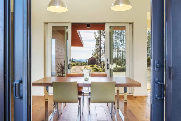 Bright dining area viewed through open dark-framed glazed doors, featuring a timber dining table with upholstered chairs, warm timber flooring, pendant lighting, and large central glazing opening to a landscaped rural-looking exterior. Pale walls and extensive glazing create an airy indoor-outdoor connection.