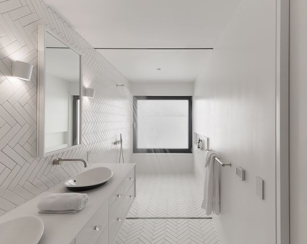 A bright, contemporary bathroom with white herringbone tilework covering the feature wall and floor. The room includes a floating white vanity with drawers, a vessel basin, wall-mounted tapware, a large frameless mirror, a walk-in shower area with a slim glass screen, and a black-framed window. Minimal chrome fixtures and neatly folded towels contribute to a clean, refined aesthetic.