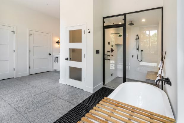 Bright contemporary bathroom with a freestanding white bathtub in the foreground, a black-framed glass shower enclosure, wall-mounted black fixtures, and a timber bath caddy. The room has pale walls, large grey floor tiles, white paneled doors, and a glass-panel internal door, creating a clean, modern, high-contrast interior.