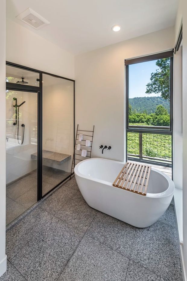 Bright contemporary bathroom featuring a freestanding white bathtub with a timber bath tray, a large frameless-style glass shower enclosure with dark metal framing, grey speckled stone-look floor tiles, white walls, black tapware, and a large window overlooking a green wooded landscape.